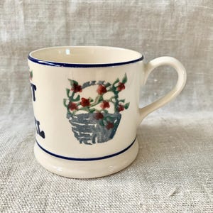 May include: Cream-colored ceramic mug with a blue rim and base. The mug features a hand-painted design of a basket of red flowers and the words "Sweet Soul" in blue. The handle is white and curved.
