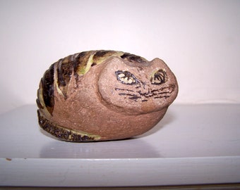 May include: A ceramic cat figurine with brown and green stripes and painted eyes. The cat is curled up in a ball.