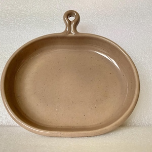 May include: A tan ceramic serving dish with an oval shape and a handle. The dish has a speckled surface and a raised rim. The handle is a small loop. The dish is likely used for serving food.
