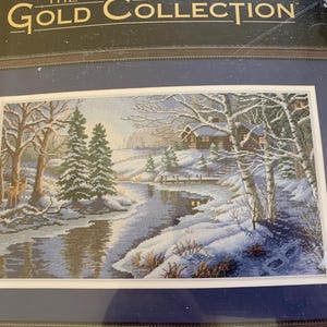 May include: Cross-stitch landscape featuring a winter scene with a snow-covered river, evergreen trees, and a cabin. The artwork is framed and set against a dark blue mat. The text "THE GOLD COLLECTION" is at the top.