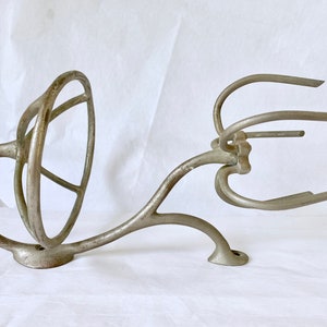 Puede incluir: Una pieza antigua de metal fundido, posiblemente de una batidora o batidor. La pieza es de color plateado con un dise&ntilde;o curvo, con un elemento circular y varios brazos curvos. El metal muestra signos de edad y desgaste.
