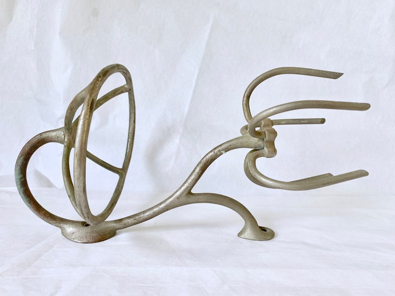 Puede incluir: Una pieza antigua de metal fundido, posiblemente de una batidora o batidor. La pieza es de color plateado con un dise&ntilde;o curvo, con un elemento circular y varios brazos curvos. El metal muestra signos de edad y desgaste.