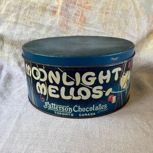 May include: A round, dark blue metal tin with a lid. The side features the text "MOONLIGHT MELLOS" in large, cream-colored letters, with "Patterson Chocolates Limited" and "TORONTO CANADA" printed below in smaller white script and block text.