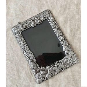 Puede incluir: Marco de fotos rectangular plateado con interior negro. El marco está decorado con detallados relieves de conejos y follaje. El marco es de metal y tiene un aspecto vintage o antiguo.