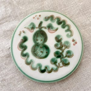 May include: A round, white ceramic coaster with a hand-painted design. The design features green and brown botanical elements, including a central pear-shaped motif and surrounding vines. The coaster has a green border and is set against a neutral linen background.
