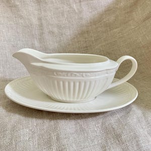 May include: A white ceramic gravy boat with a matching saucer. The gravy boat has a spout and handle, and both pieces feature a ribbed design. The set is displayed on a neutral-colored linen surface.