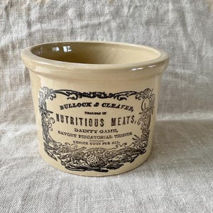 May include: A cream-colored stoneware crock with a black label featuring the text "BULLOCK & CLEAVER, NUTRITIOUS MEATS, DAINTY GAME, SAVORY PISCATORIAL TIDBITS, CHOICE CUTS FOR ALL." The antique-style container has a round shape.