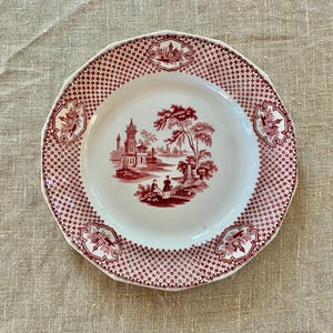 Puede incluir: Plato de cerámica blanca con un patrón rojo y blanco. El borde tiene un borde festoneado y un patrón de cuadros rojos. El centro del plato presenta una escena de paisaje rojo con edificios y árboles. Elementos florales decorativos se colocan alrededor del borde.
