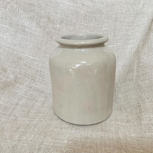May include: A small, off-white ceramic jar with a rounded body and a slightly flared rim. The jar has a smooth, glossy finish and sits on a neutral-colored linen surface. The jar is likely for storage or decorative purposes.