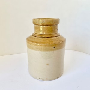May include: A vintage ceramic jar with a light brown top and a white bottom. The jar has a simple, cylindrical shape and a smooth, glazed finish.