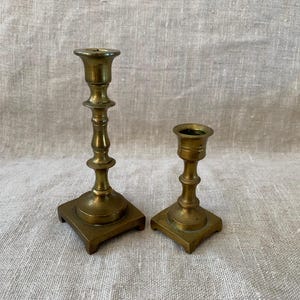 May include: Two antique brass candlesticks. The taller candlestick has a fluted stem and a square base. The shorter candlestick has a similar design. Both are in a warm, golden hue, suggesting age and use. The background is a neutral linen fabric.