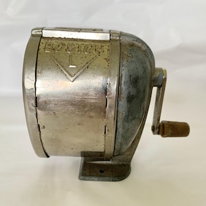 May include: Vintage silver-toned pencil sharpener with the word "BOSTON" embossed on the front. The sharpener has a hand crank and a rectangular base. The metal shows signs of age and wear, with a slightly tarnished appearance.