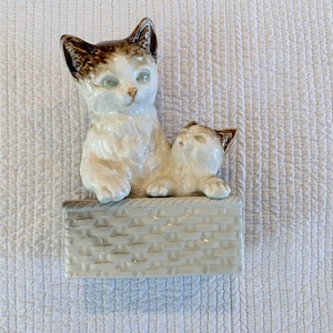 May include: A white ceramic figurine of two cats sitting in a basket. The larger cat is brown and white with green eyes, and the smaller cat is white with brown markings. The basket is decorated with a woven pattern.