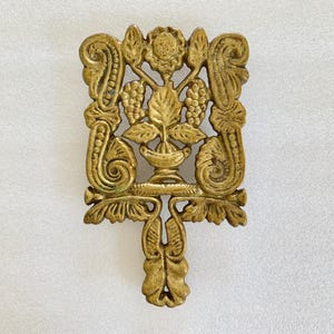 May include: Ornate, antique brass trivet with a detailed floral and grape design. The trivet features a central floral motif, flanked by grape clusters and scrolling embellishments. The handle extends from the bottom, adding to its decorative appeal.