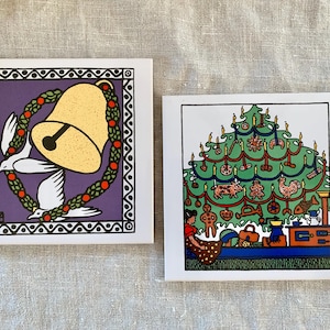 May include: Two square Christmas-themed art prints. One features a gold bell with a black clapper, surrounded by a wreath and doves, set against a purple background. The other depicts a decorated Christmas tree with ornaments and a woman.