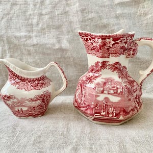 May include: Two antique cream pitchers with a red and white toile pattern. The larger pitcher has a detailed landscape scene and a decorative handle. The smaller pitcher has a similar design. Both are on a neutral linen surface.