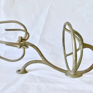 Puede incluir: Un utensilio de cocina antiguo de hierro fundido, probablemente un batidor o componente de mezclador. La pieza tiene un dise&ntilde;o complejo y calado con brazos curvos y un elemento circular. El metal tiene un aspecto envejecido, de color gris plateado apagado.