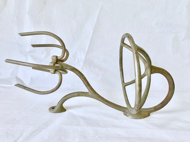 Puede incluir: Un utensilio de cocina antiguo de hierro fundido, probablemente un batidor o componente de mezclador. La pieza tiene un dise&ntilde;o complejo y calado con brazos curvos y un elemento circular. El metal tiene un aspecto envejecido, de color gris plateado apagado.