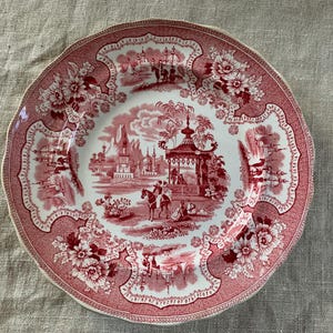 Puede incluir: Plato redondo de estilo antiguo, con borde festoneado. El diseño rojo y blanco presenta motivos florales y una escena central con figuras, edificios y árboles. Un artículo decorativo.