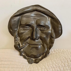May include: A gray, sculpted face of a man with a beard and a pipe. The man is wearing a hat and has a serious expression.