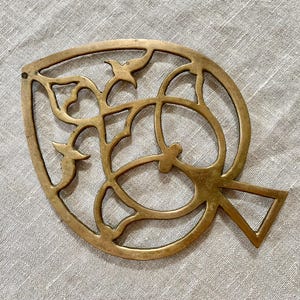 May include: A decorative brass leaf-shaped trivet with an openwork design. The trivet features a leaf shape with intricate cutouts of floral and geometric patterns. The brass has a warm, golden hue, and the trivet is placed on a neutral linen surface.