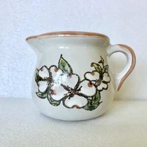 May include: Cream-colored ceramic creamer with a brown rim and handle. The creamer is decorated with white flowers with brown centers and green leaves. The creamer has a small spout for pouring.