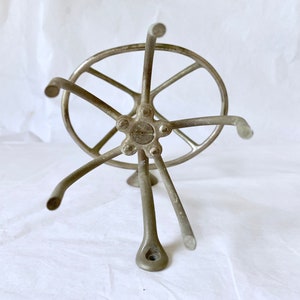 Puede incluir: Un ovillador de hilo plateado vintage, con un cubo central con brazos radiales y un anillo exterior circular. El ovillador tiene una base de tr&iacute;pode y est&aacute; dise&ntilde;ado para sujetar el hilo para enrollar. El metal tiene un aspecto ligeramente envejecido.
