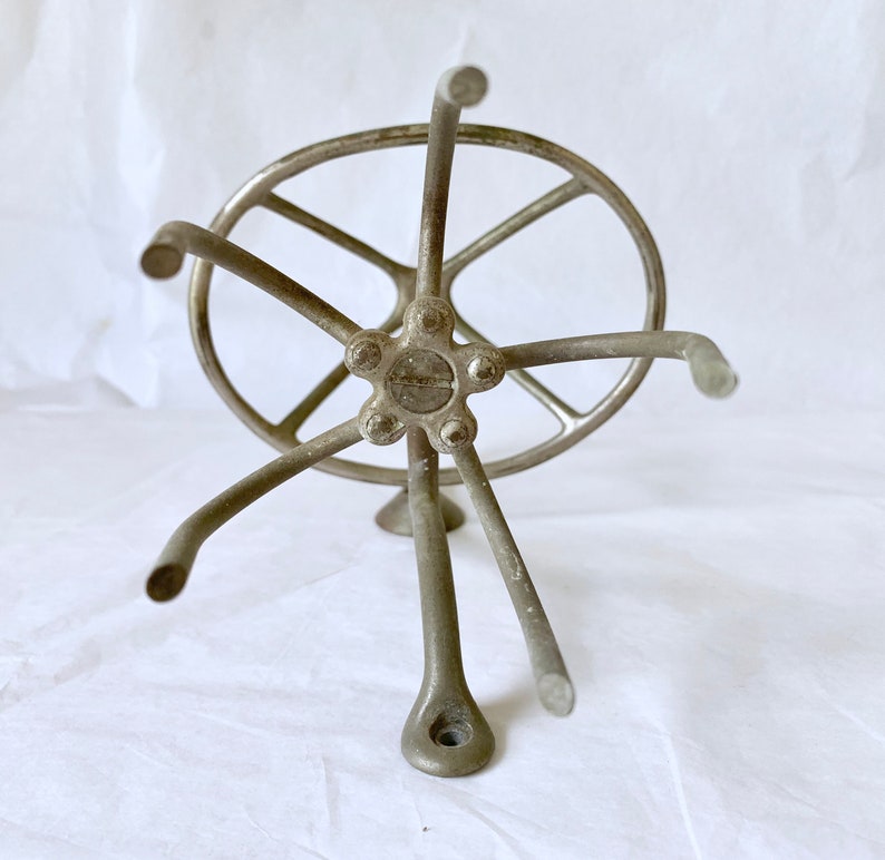 Puede incluir: Un ovillador de hilo plateado vintage, con un cubo central con brazos radiales y un anillo exterior circular. El ovillador tiene una base de tr&iacute;pode y est&aacute; dise&ntilde;ado para sujetar el hilo para enrollar. El metal tiene un aspecto ligeramente envejecido.