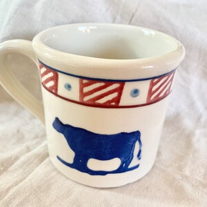 May include: White ceramic mug with a blue cow silhouette and a decorative band of red and blue geometric patterns. The mug has a curved handle and a cream-colored rim, perfect for enjoying your favorite beverage.