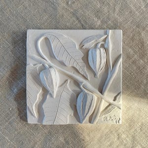May include: A square, off-white relief sculpture featuring a botanical design. The artwork depicts stylized leaves, branches, and seed pods. The piece is signed and dated. The overall theme is nature and botanical art.