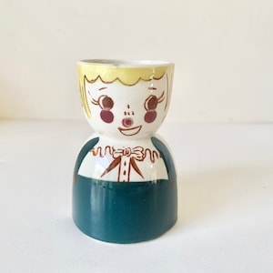 May include: A vintage ceramic egg cup shaped like a smiling child. The egg cup features a white face with yellow hair, rosy cheeks, and a teal blue body. The design includes a brown bow tie and painted details.