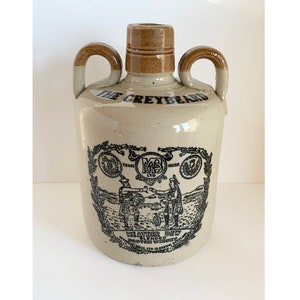 May include: A ceramic jug with two handles, featuring a black and white illustration of two men in kilts shaking hands. The jug is labeled "The Greybeard Weather Dew Blended Whisky" and "M.B.S. Ltd."