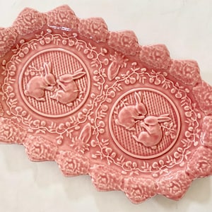 May include: A pink ceramic serving dish with a scalloped edge and a raised design of two bunnies in each of two circular sections. The dish is decorated with a floral pattern.