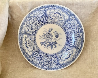 May include: A round, white ceramic plate with a blue floral design. The plate features a central floral motif surrounded by ornate patterns and a detailed border. The design includes roses and other flowers.