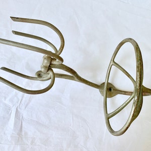 Puede incluir: Un utensilio de cocina antiguo, de color plateado, posiblemente un batidor o un accesorio de batidora. Presenta un mango circular con una serie de radios curvos y un extremo con p&uacute;as. El metal muestra signos de edad y uso.
