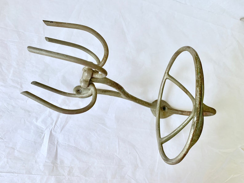 Puede incluir: Un utensilio de cocina antiguo, de color plateado, posiblemente un batidor o un accesorio de batidora. Presenta un mango circular con una serie de radios curvos y un extremo con p&uacute;as. El metal muestra signos de edad y uso.