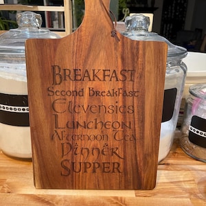 May include: A wooden cutting board with a brown leather handle. The board has the words "Breakfast", "Second Breakfast", "Elevensies", "Luncheon", "Afternoon Tea", "Dinner", and "Supper" engraved on it.