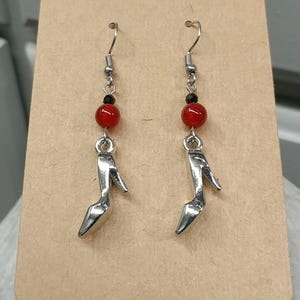 May include: A pair of dangle earrings featuring silver-toned high-heeled shoe charms. Each earring includes a small black bead and a larger round red bead suspended from a silver-toned hook wire, creating a vertical design with a metallic finish.