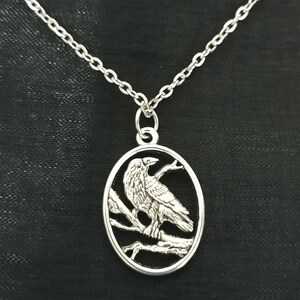 May include: Silver-toned raven pendant necklace. The oval pendant features a detailed raven perched on a branch within an oval frame. The necklace has a delicate chain and a simple clasp. The raven is facing left.