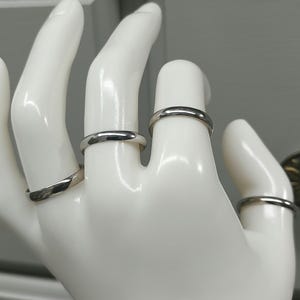 May include: Four polished silver-toned metal bands worn on the fingers of a white mannequin hand. Each ring features a smooth, rounded, and minimalist design with a reflective surface, showcasing a consistent metallic finish across all four pieces.