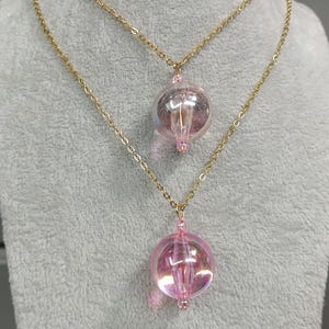May include: Two gold-coloured chain necklaces. Each necklace has a pendant with a pink, iridescent, spherical bead. The beads have a clear, vertical, rod-shaped element. The necklaces are displayed on a grey surface.