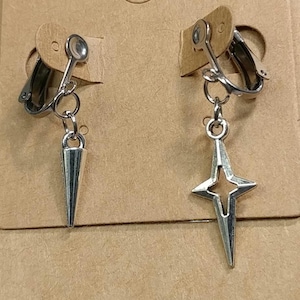 Op de afbeelding: Zilverkleurige clip-oorbellen. Eén oorbel heeft een kegelvormige hanger, de andere een vierpuntig sterontwerp. De oorbellen worden gepresenteerd op een bruine kartonnen achterkant.