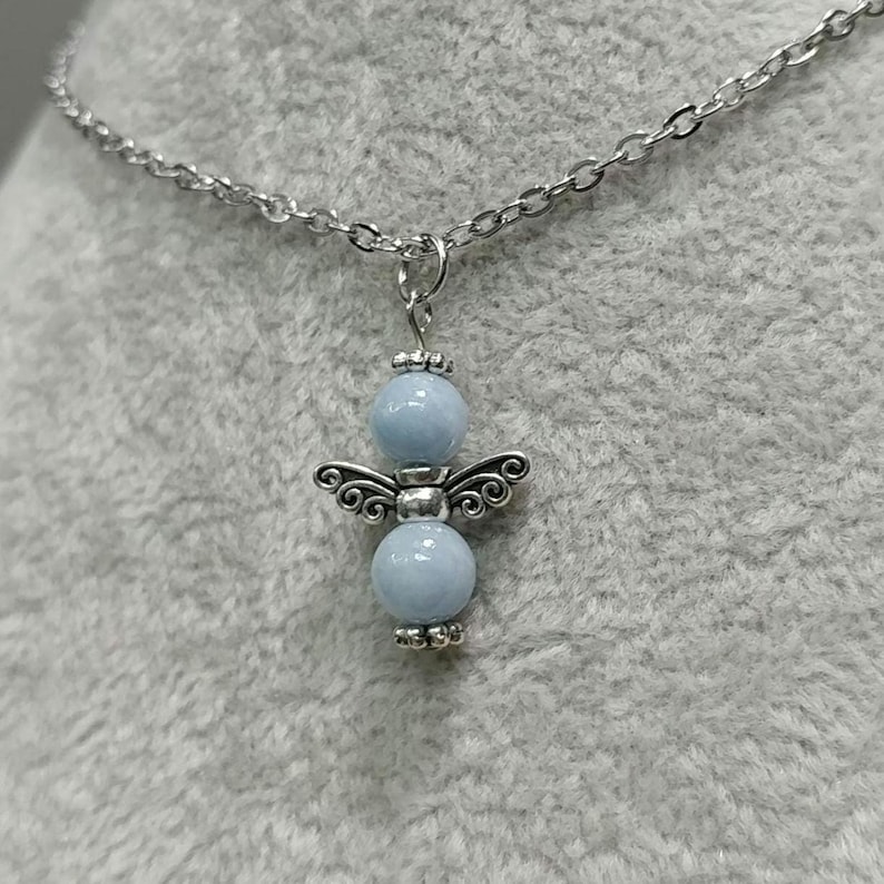 May include: A silver-toned necklace featuring a pendant shaped like an angel. The angel pendant has a light blue bead for the head and body, with silver-toned wings and decorative accents. The necklace chain is also silver-toned.