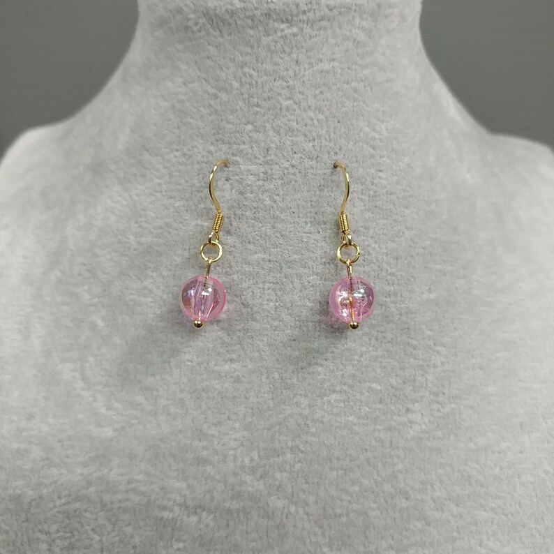 May include: A pair of gold dangle earrings with pink glass beads. The beads are round and have a slight shimmer.