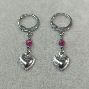 May include: A pair of silver-toned dangle earrings. Each earring features a heart-shaped charm with a beaded edge, suspended from a pink bead and a lever-back closure. The earrings are displayed against a light gray background.