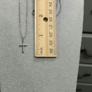 Pode incluir: Um colar de corrente de prata com um pequeno pingente de cruz de prata. O colar &eacute; medido com uma r&eacute;gua, mostrando que tem aproximadamente 50 cm de comprimento.