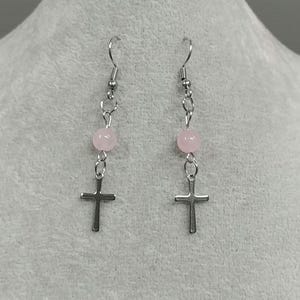 Puede incluir: Un par de pendientes de plata con una cuenta rosa y un dije de cruz de plata.