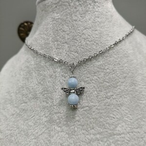 May include: A silver-toned necklace featuring a pendant shaped like an angel. The angel pendant has two light blue beads for the body and head, with silver-toned wings. The necklace is displayed on a gray surface.