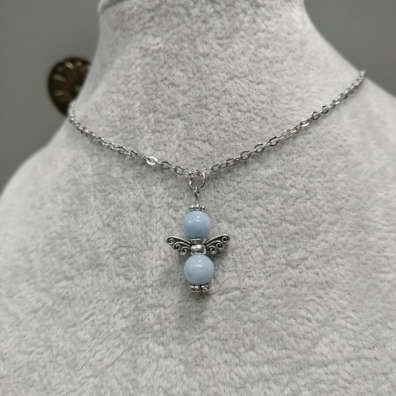May include: A silver-toned necklace featuring a pendant shaped like an angel. The angel pendant has two light blue beads for the body and head, with silver-toned wings. The necklace is displayed on a gray surface.