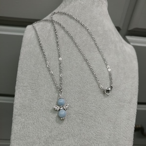 May include: A silver-toned necklace featuring a pendant shaped like an angel. The angel pendant has a light blue bead body and decorative wings. The necklace is displayed on a gray surface.